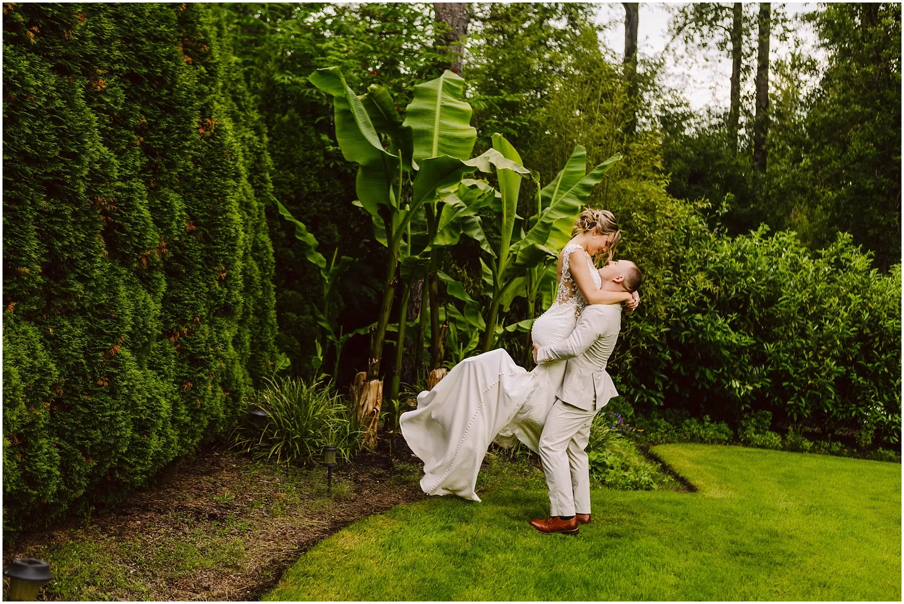 Snohomish Wedding Photographer GSquared Weddings In a lush Snohomish garden, a groom in a light suit lifts the bride in her white dress amid vibrant green foliage and banana plants. Their joy is palpable as she gazes at him lovingly while being held. Serving Seattle, Snohomish and Orlando
