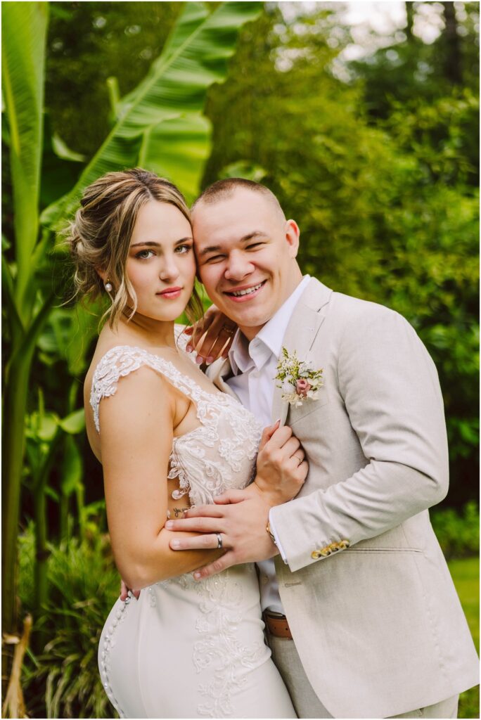 Snohomish Wedding Photographer GSquared Weddings A couple in wedding attire poses outdoors in Snohomish The woman wears a lace wedding dress and the man is in a light beige suit with a boutonniere They are surrounded by lush green foliage including large banana leaves and both are smiling Serving Seattle Snohomish and Washington State Snohomish Wedding Photographer GSquared Weddings A couple in wedding attire poses outdoors in Snohomish. The woman wears a lace wedding dress, and the man is in a light beige suit with a boutonniere. They are surrounded by lush green foliage, including large banana leaves, and both are smiling. Serving Seattle, Snohomish and Washington State