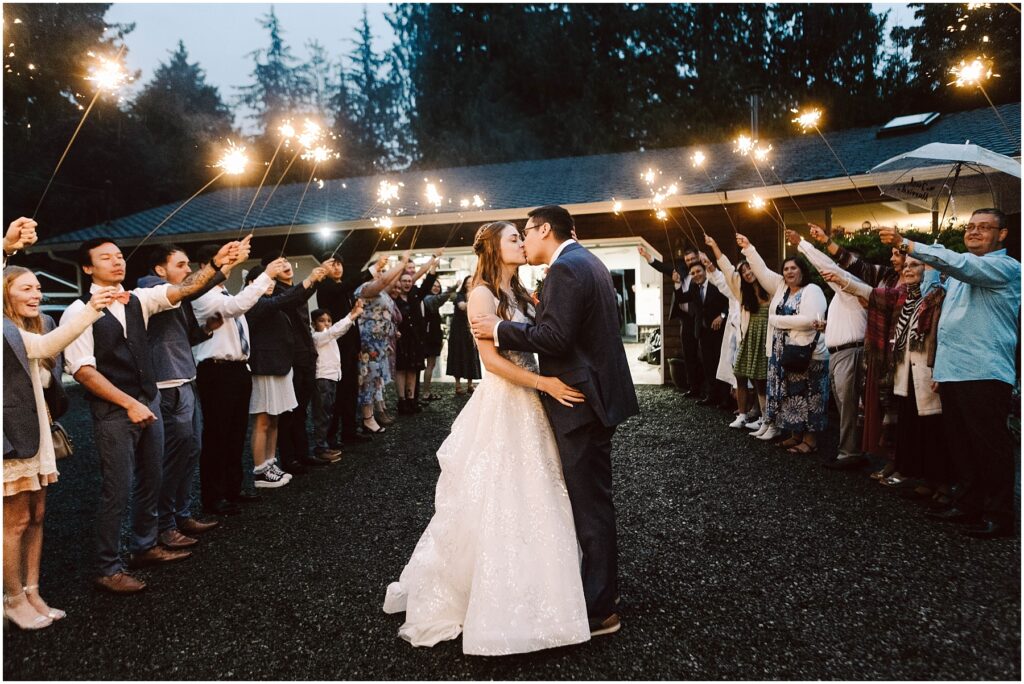 Snohomish wedding photographer gsquared weddings a bride and groom share a kiss under the night sky during their backyard wedding surrounded by guests holding sparklers The scene is festive with an arch of light formed by the guests and a backdrop of trees and a lit building Serving seattle snohomish and washington state