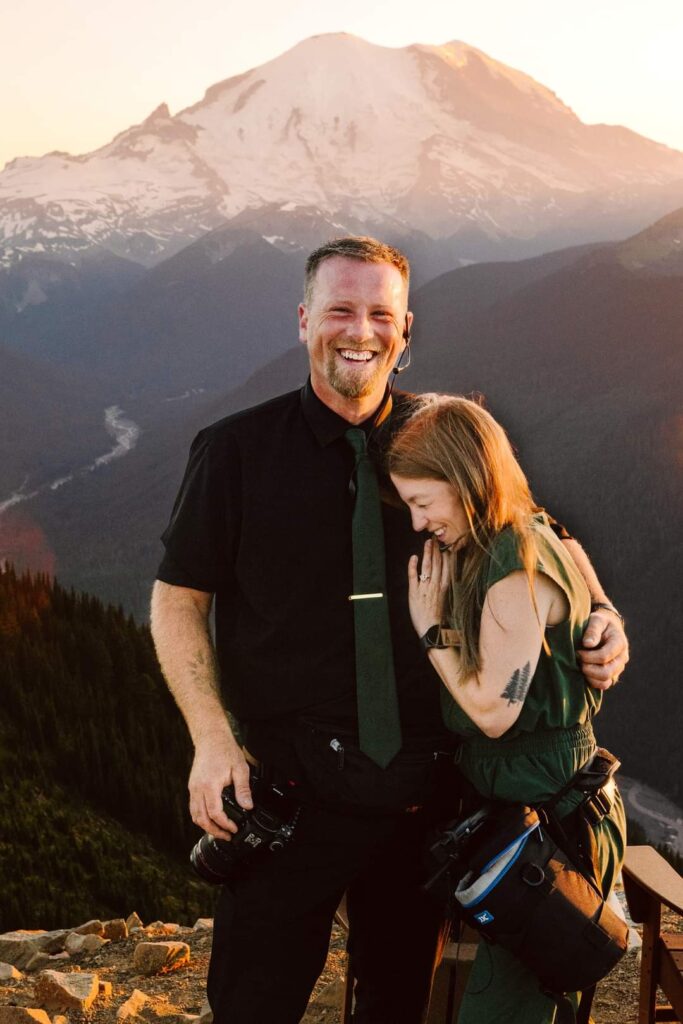 GSquared Weddings Photography A man and woman stand close together smiling and laughing on a mountain overlook at sunset Snowcapped peaks and forested hills create a scenic backgroundperfect inspiration for an About page designed in WordPress with Elementor Both wear camera gear suggesting they are photographers GSquared Weddings | | Serving Snohomish Seattle Beyond