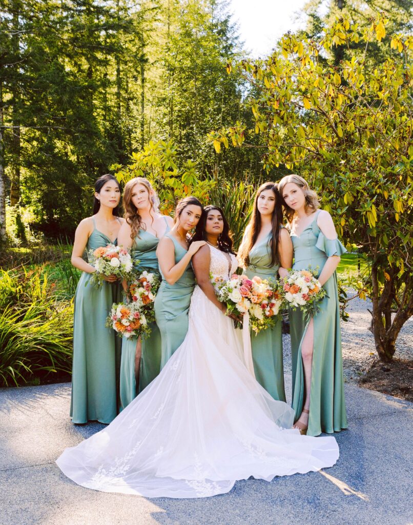 GSquared Weddings Photography A bride in a white gown stands outdoors with five bridesmaids in sage green dresses each holding bouquets surrounded by lush greenery and sunlightperfect inspiration for showcasing wedding designs on your WordPress or Elementor services website GSquared Weddings | | Serving Snohomish Seattle Beyond GSquared Weddings Photography bridesmaids in sage green dresses