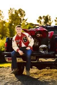 Smiling young man in a letterman jacket sitting on a pickup tailgate beside a red atv with string lights overhead