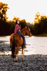 Woman in a red patterned shirt and jeans riding a brown horse along a riverbank at sunset