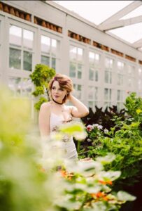 Woman in a light dress in a sunlit greenhouse with her hand touching her hair