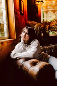 Woman in a white sweater sitting on a brown leather couch by a window with her chin resting on her hand