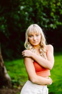 Young woman with long blonde hair in a sleeveless orange top and light pants one arm crossed over her body
