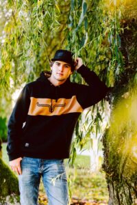 Young man in a black and tan hoodie jeans and black cap leaning against a tree