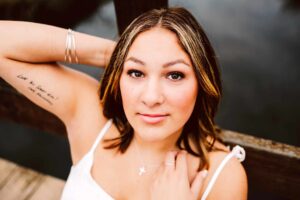 Young woman with shoulder length hair and subtle blonde highlights in a white top with silver bracelets and a cross necklace arm behind her head tattoo on her upper arm