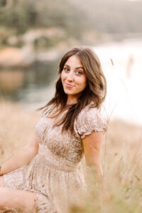 Senior with long brown hair in a light floral dress sitting on dry grass near a lake and forest smiling softly