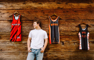 Senior in a white t shirt and jeans standing in front of a wooden wall hung with sports jerseys