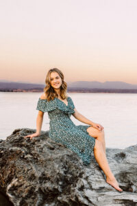 Smiling senior in a green floral dress sitting barefoot on a large rock by a calm lake at sunset