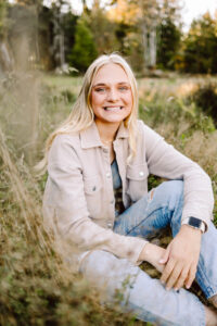 Smiling senior with long blonde hair in a light jacket and ripped jeans sitting in a field
