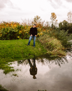 Senior in a black sweater jeans and cap standing on grass beside a pond with their reflection in the water