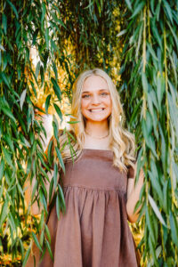 Smiling senior with long blonde hair in a brown dress standing among hanging willow branches