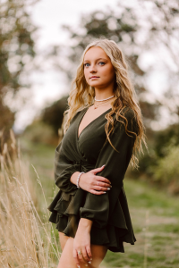 Senior with long wavy blonde hair in a dark green wrap dress standing in tall grass