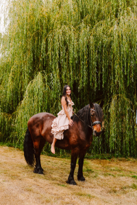Senior in a beige dress sitting barefoot on a large brown horse with tall willow trees behind her