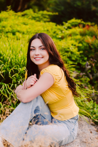 Smiling senior in a yellow top and light blue jeans sitting on a rock surrounded by lush green plants