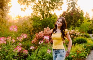 Smiling senior with long dark hair in a yellow top and jeans standing among blooming pink flowers in a garden at sunset