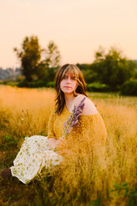 Senior in a yellow sweater and floral skirt sitting in tall grass at sunset holding a bunch of purple flowers