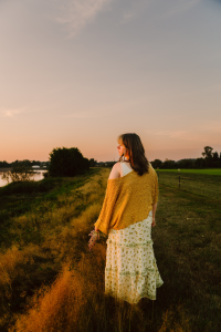 Senior in a floral dress and yellow sweater standing in tall grass near a river at sunset looking into the distance