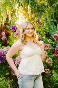 Senior with blonde hair in a white camisole and jeans smiling in a garden full of hydrangeas