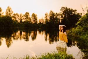 Senior in a yellow top and floral skirt standing knee deep in a lake at sunset with an orange sky reflected on the water