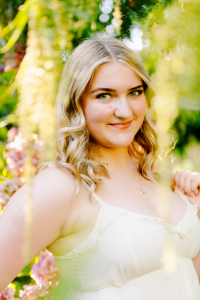 Senior with wavy blonde hair in a white dress and cross necklace smiling among green foliage and pink flowers