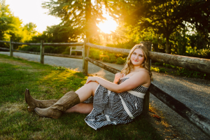 Senior in a checkered dress and cowboy boots sitting on the grass by a wooden fence at sunset