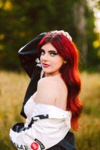 Senior with long wavy red hair posing in a field wearing a white top with a black and white jacket draped over her shoulders