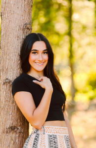 Senior with long dark hair in a black crop top and patterned skirt leaning against a tree and smiling