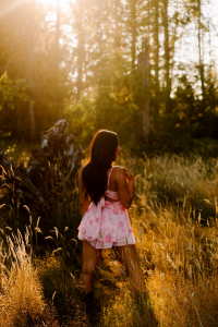 Senior in a pink floral dress standing in a sunlit grassy field with trees and golden light streaming through the leaves