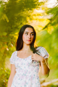 Senior in a light floral dress looking to the side surrounded by lush green foliage and warm sunlight