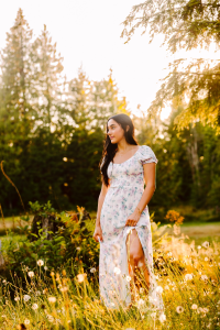 Senior in a light floral dress standing in a sunlit meadow of wildflowers and tall grass with a forest behind her