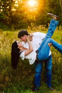 Couple laughing as a man in jeans and a white shirt lifts and embraces a smiling woman in a sunlit field of tall grass