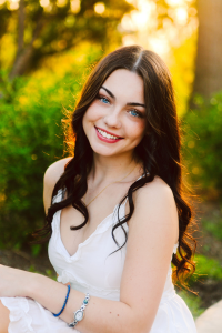 Senior with long wavy dark hair and blue eyes in a white dress smiling while sitting outdoors in sunlight