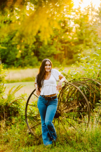 Smiling senior in a white top and blue jeans posing in front of an old wagon wheel in a sunlit field