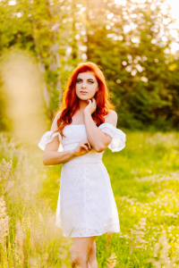 Senior with long red hair in a white off the shoulder dress standing in a sunlit grassy field