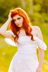 Senior with long red hair in a white off the shoulder dress touching her hair against a soft green and yellow blurred background