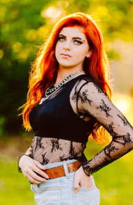 Senior with bright red hair in a sheer black lace top and high waisted jeans standing outdoors in sunlight