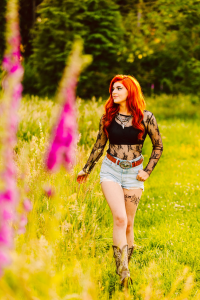 Senior with long red hair in a black lace top denim shorts and cowboy boots walking through a sunlit field with pink flowers