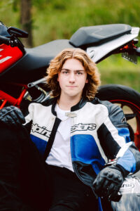 Senior with wavy shoulder length hair in a blue black and white motorcycle jacket sitting beside a black and red motorcycle