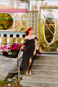 Red haired senior in a black polka dot off the shoulder dress standing on the steps of a retro diner among potted flowers