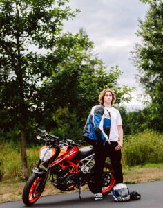 Senior standing next to an orange and white motorcycle on a road holding a blue and white jacket and helmet