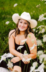 Senior in a black dress and white cowboy hat sitting in a field of wildflowers smiling softly