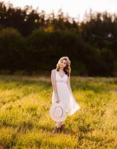 Smiling woman in a white dress holding a hat in a sunlit field of tall grass