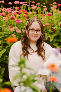 Person with long brown hair glasses and a white sweater among tall pink flowers