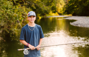 Young person in a blue shirt and cap standing in a shallow river with a fishing rod