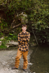 Young person in a plaid shirt and brown pants on a rocky path by the water arms crossed