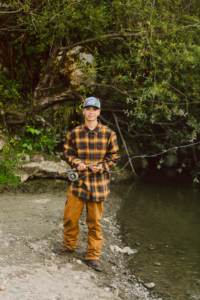Young person in a plaid shirt tan pants and cap on a riverbank holding a fishing rod