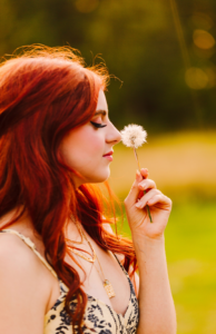 Young woman with long red hair holding a dandelion to her face eyes closed in a grassy field